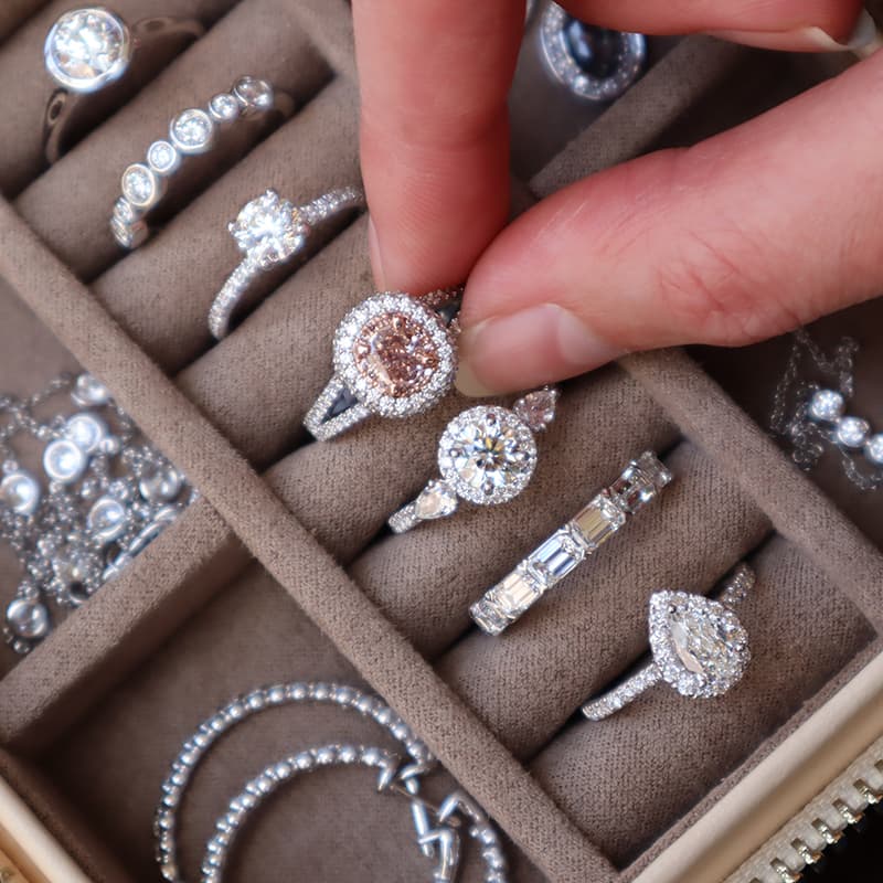 A hand lifting a pink diamond halo ring from a velvet tray of diamond engagement rings and eternity bands.