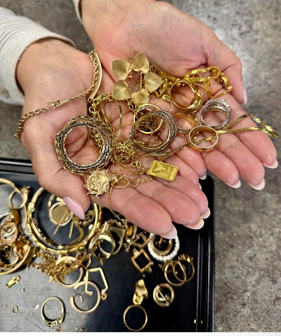Two hands holding a pile of gold chains, rings, charms and a small gold bar over a tray of more gold jewelry.