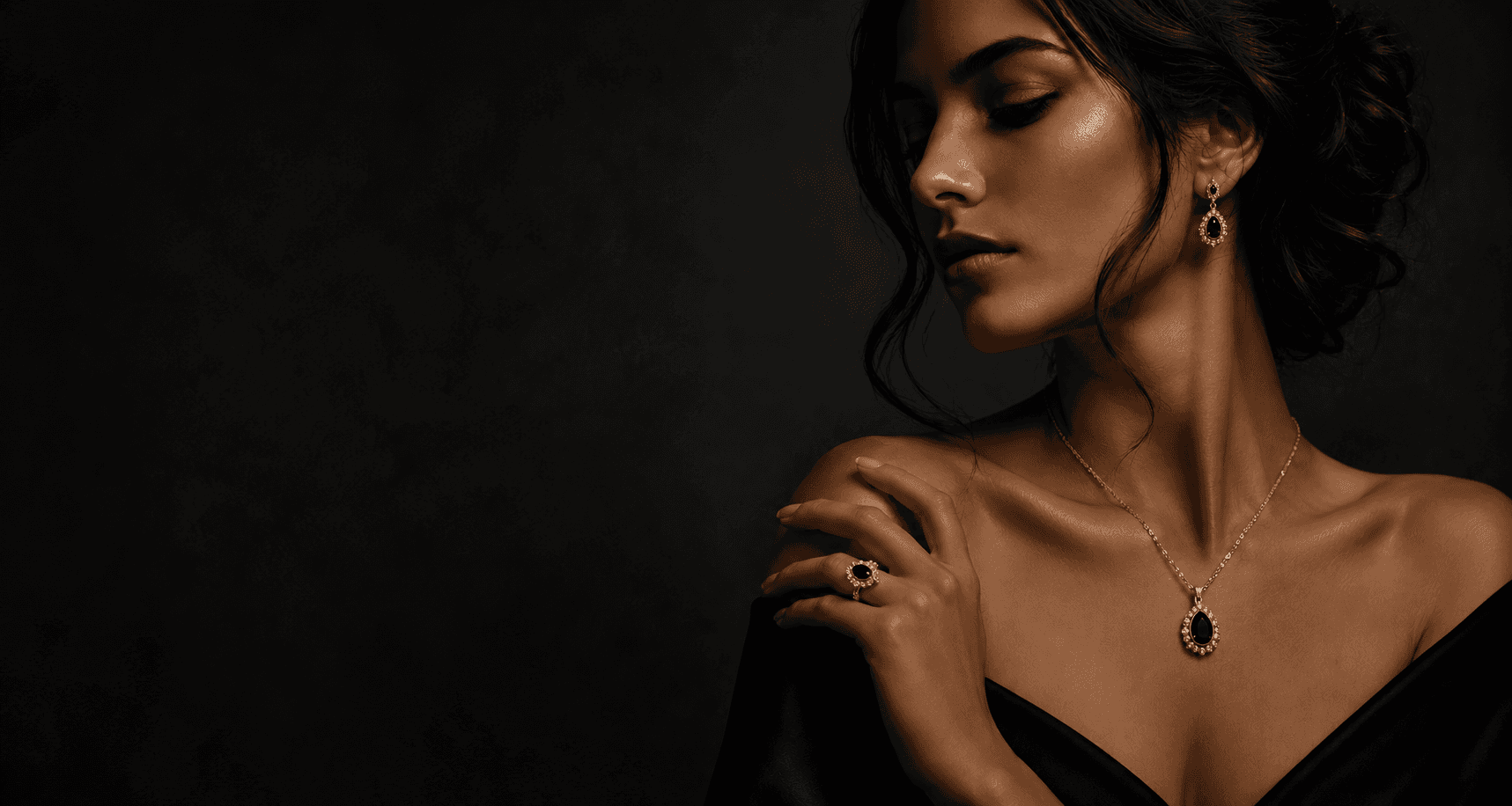 A woman wearing a gold pendant necklace, matching gold drop earrings and a gold ring, each set with a dark faceted gemstone.