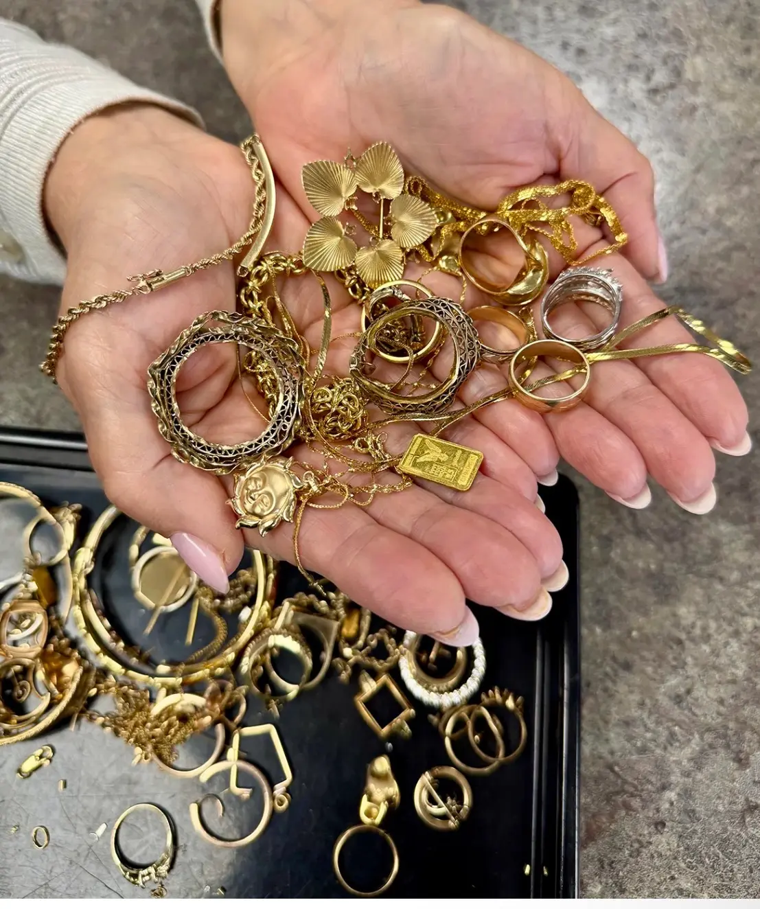Hands holding gold chains, rings, charms and a small gold bar over a tray of gold jewelry.